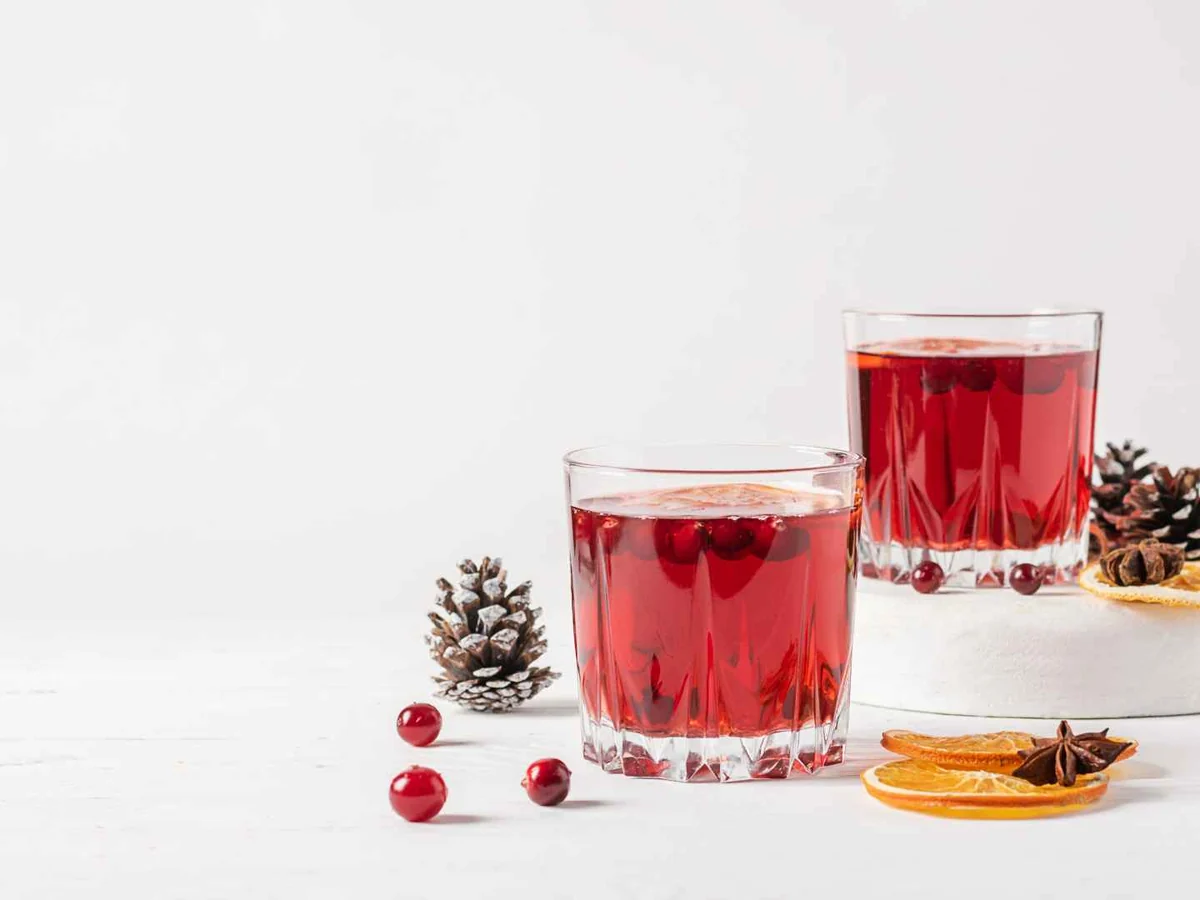 Two red cranberry drinks beside cranberries, citrus slices, and pine cones