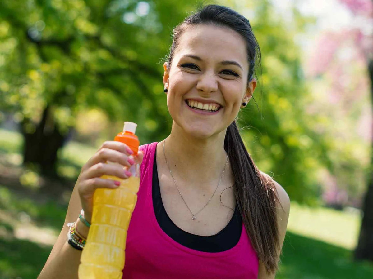A smiling woman holding an orange sports drink bottle outdoors