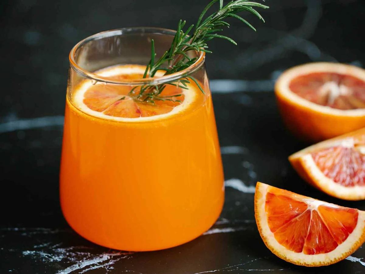 A glass of orange drink beside halved blood oranges on a dark surface