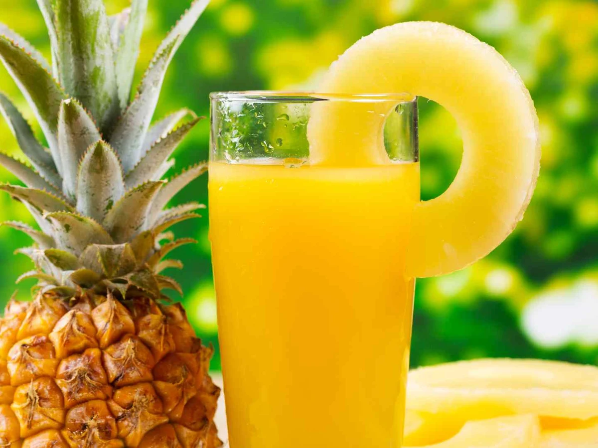 A pineapple beside a glass of juice garnished with a pineapple ring.