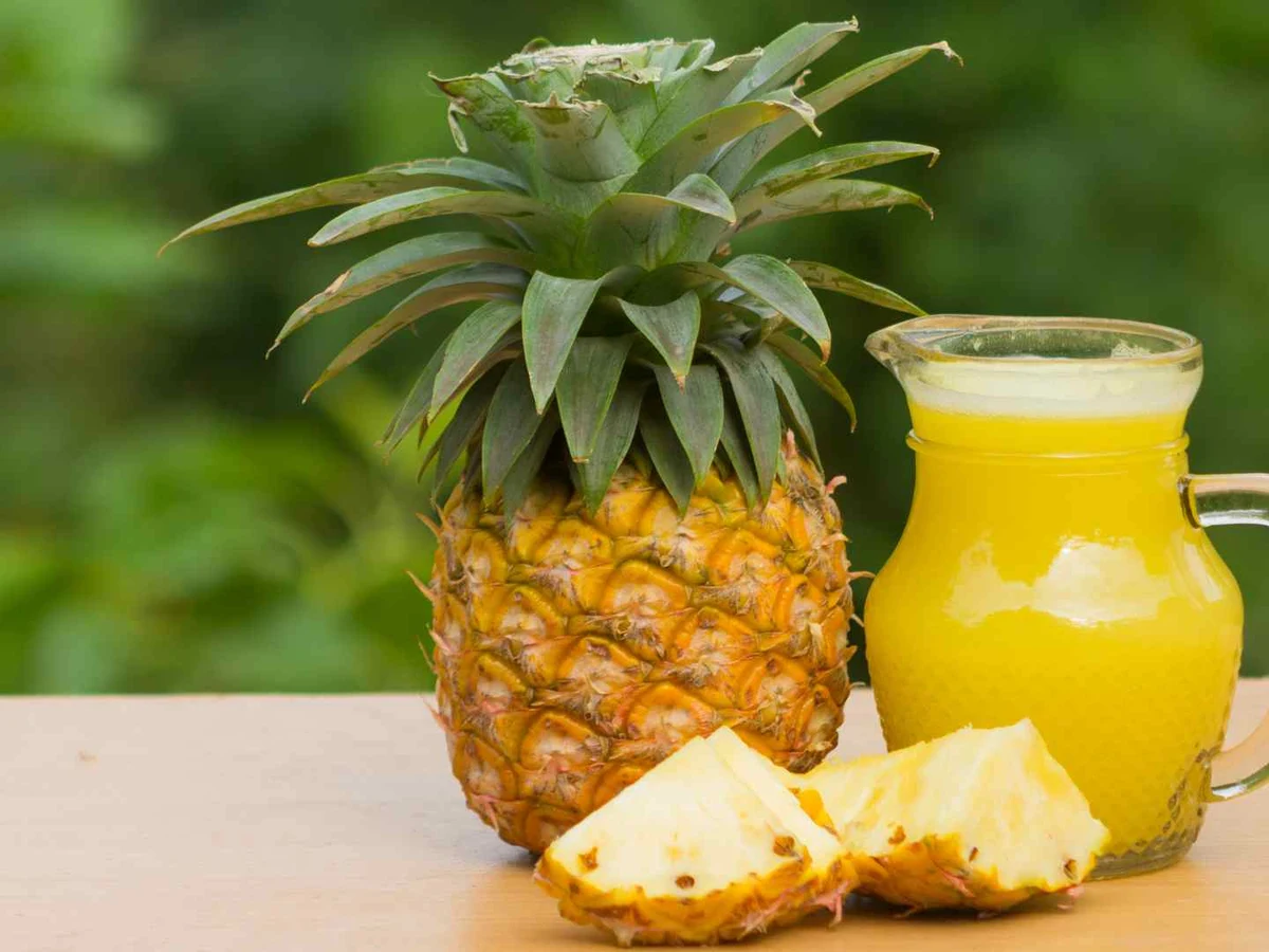 A whole pineapple with cut wedges beside a clear pitcher of yellow juice.