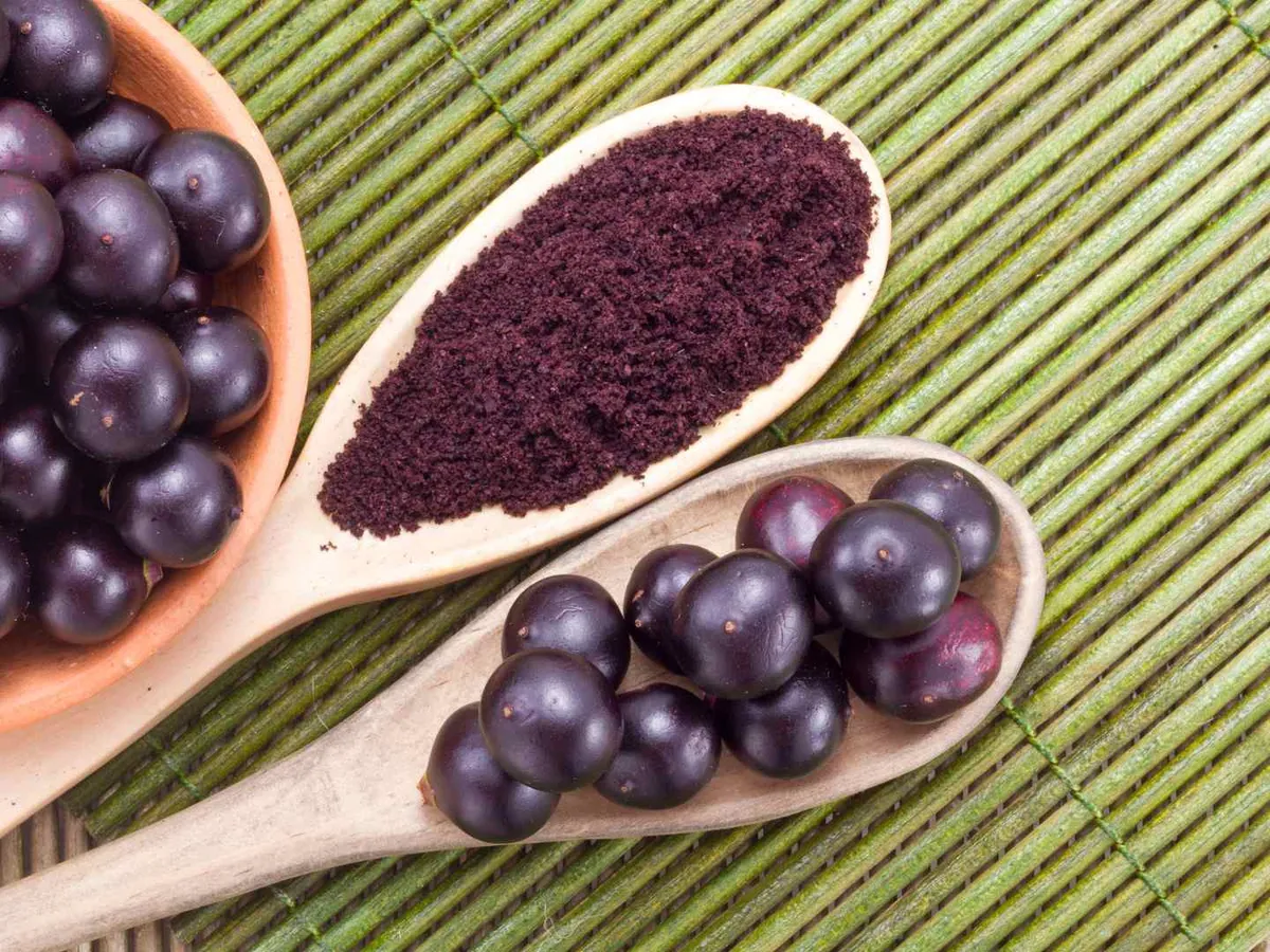 Dark purple acai powder in a wooden spoon beside round acai berries.