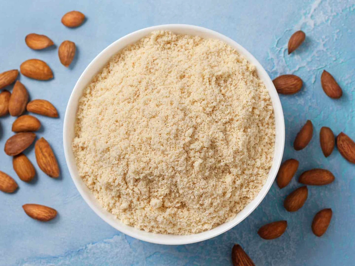 A white bowl of almond flour with whole almonds on a blue surface