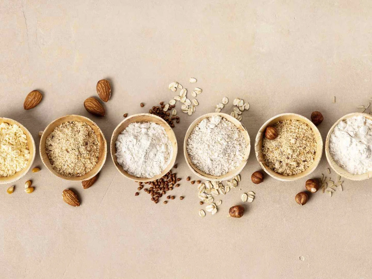 Several bowls of different flours with grains and nuts on a beige surface
