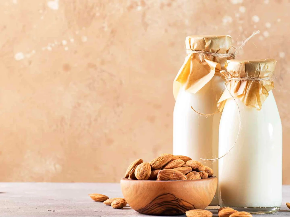Two milk bottles, a bowl of almonds, and scattered almonds on a peach backdrop
