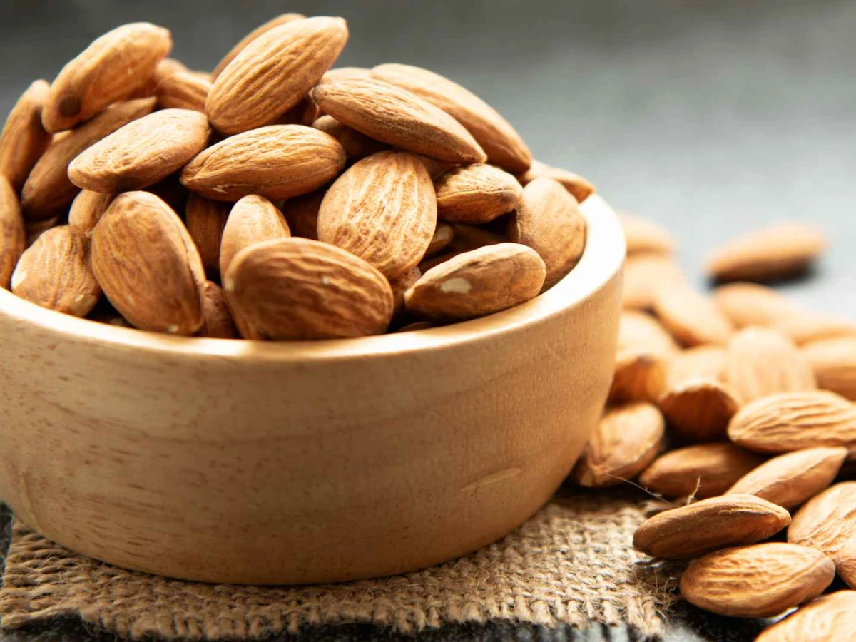 Whole almonds piled in and around a small wooden bowl.