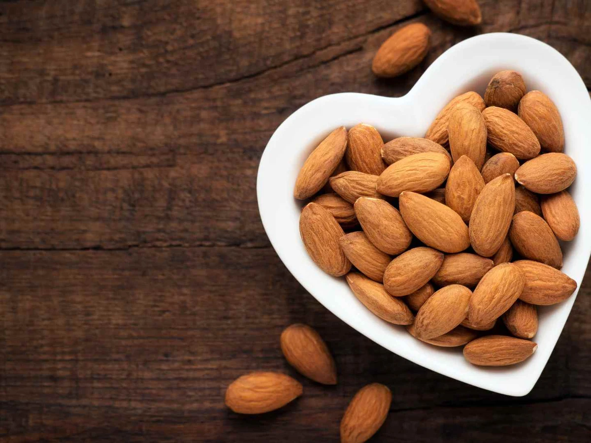Whole almonds in a white heart-shaped bowl on a dark wooden table.