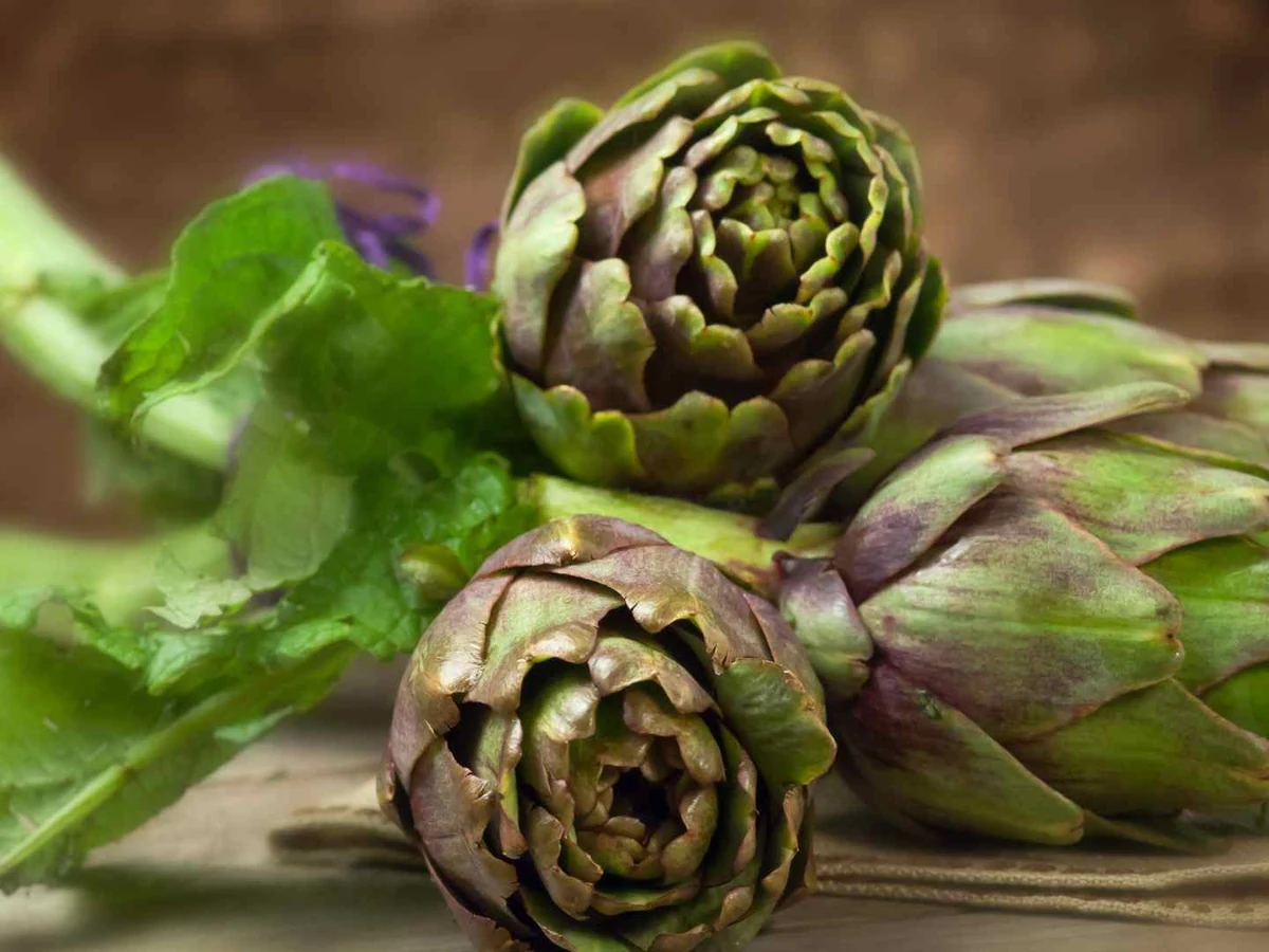 Three whole artichokes with stems resting on a rustic surface.