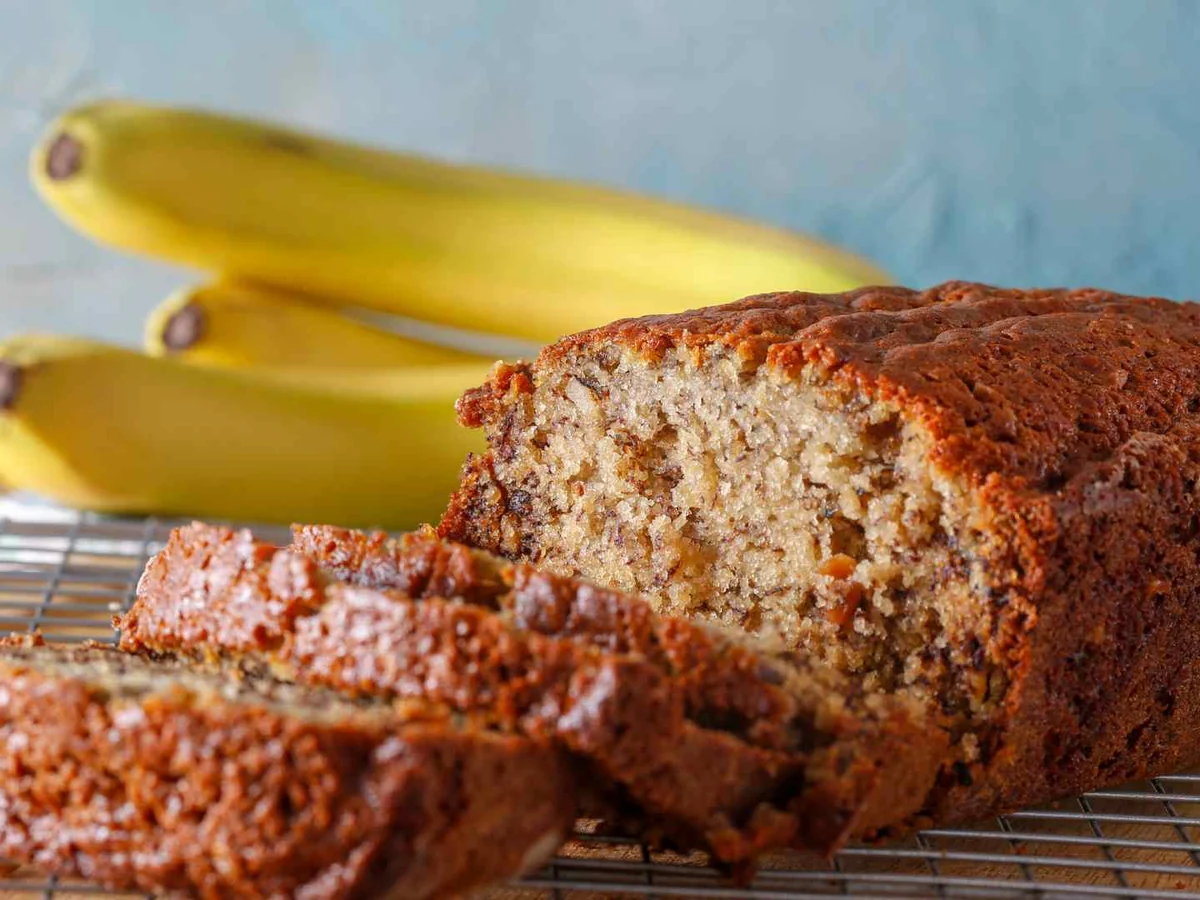 A sliced loaf of banana bread on a cooling rack with bananas behind it