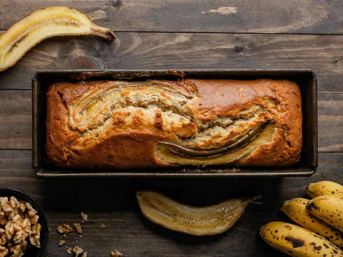 A loaf of banana bread in a pan with bananas and walnuts around it