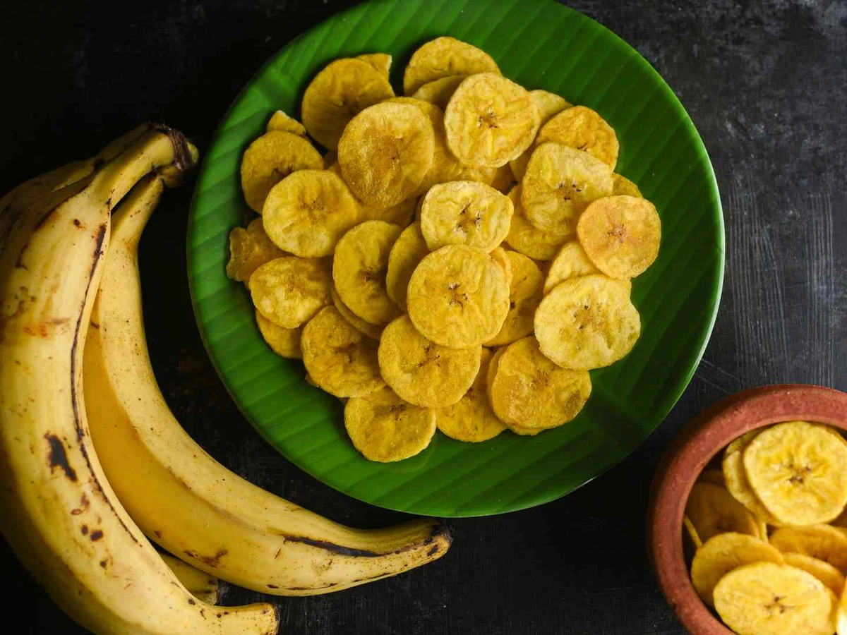 Banana chips on a green leaf beside fresh bananas on a dark background.