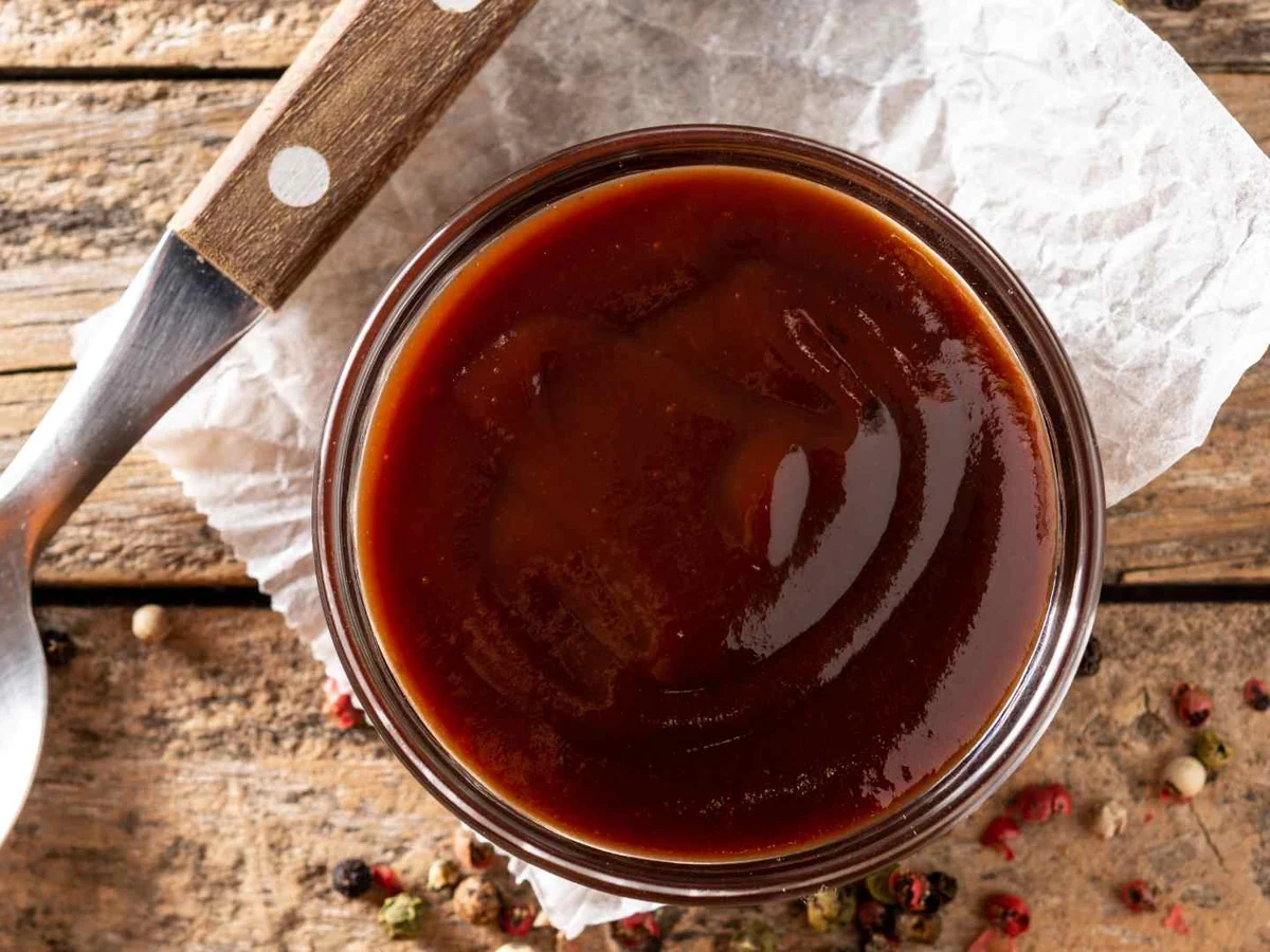 A bowl of dark barbecue sauce beside a knife on a wooden table