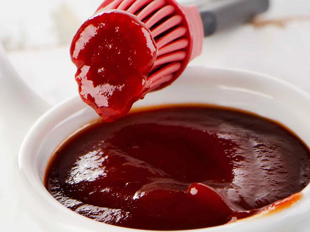 A basting brush lifting red barbecue sauce from a white bowl