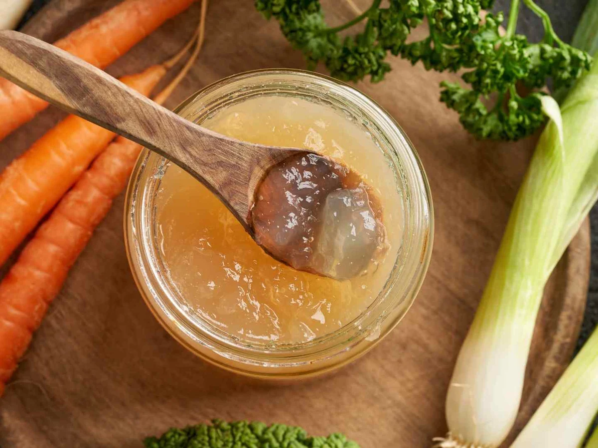 A jar of beef broth beside carrots, leek, and herbs