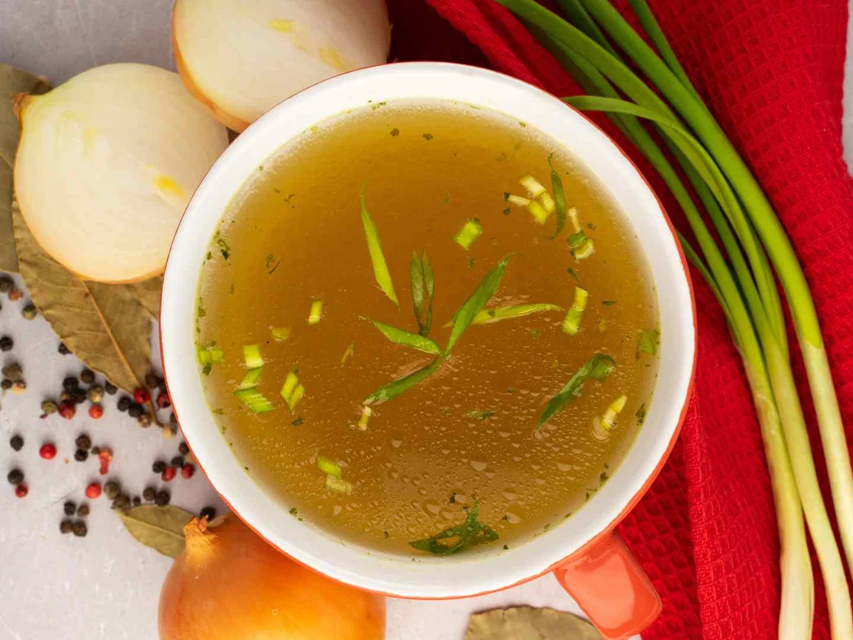 A bowl of beef broth with onion, carrot, and herbs nearby