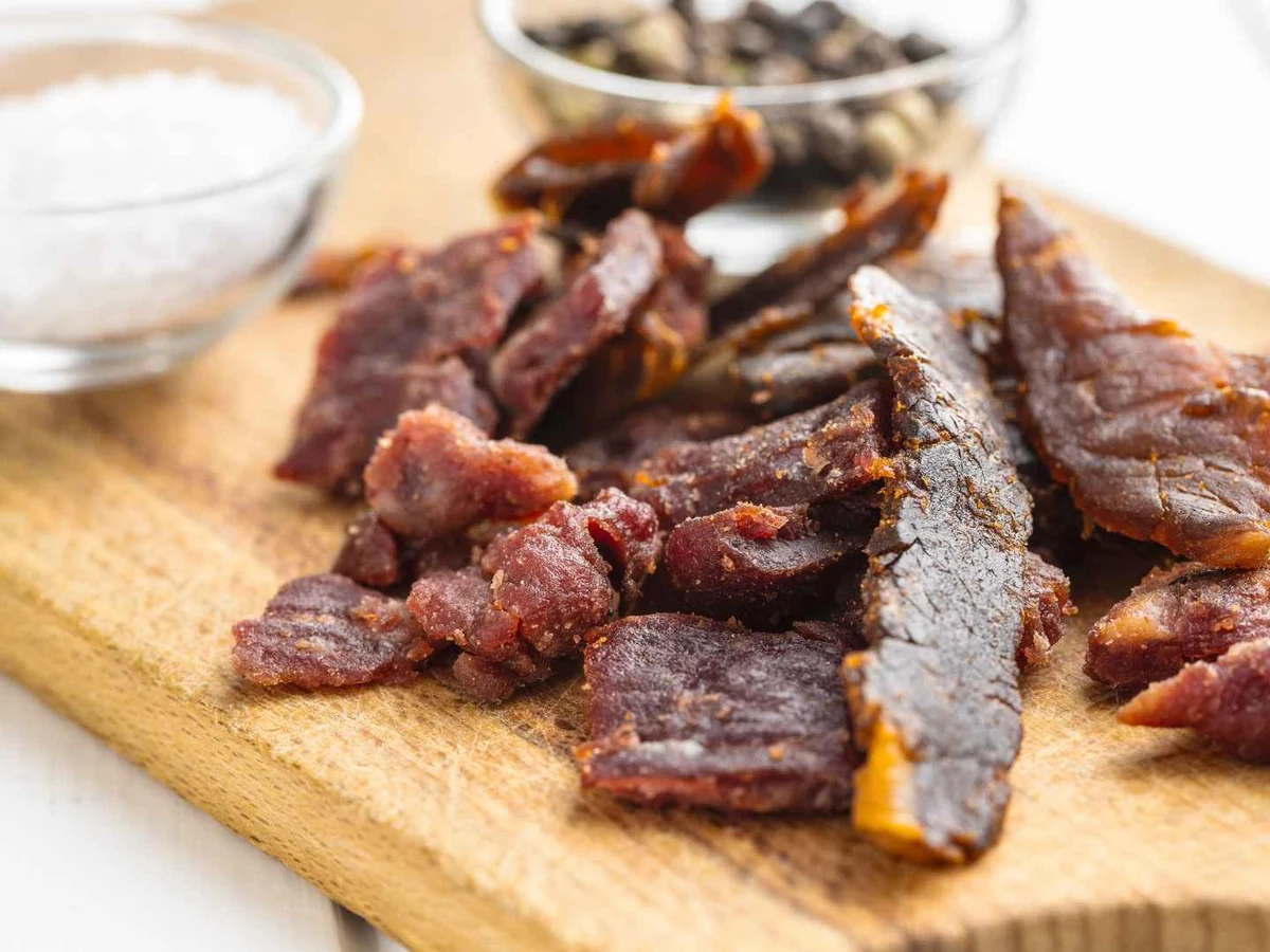 Mixed strips of dark and reddish beef jerky on a cutting board beside salt.