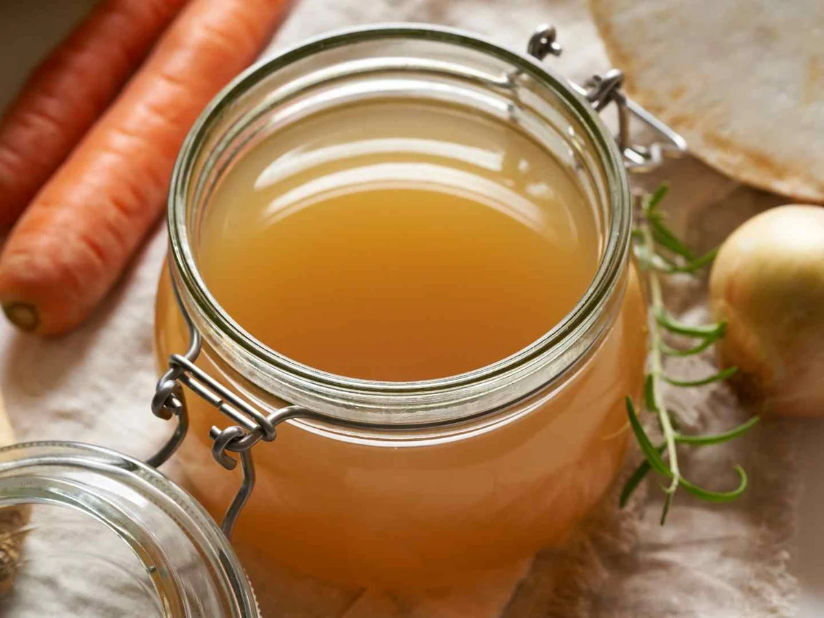 A jar of golden broth beside carrots, onion, garlic, bread, and rosemary.