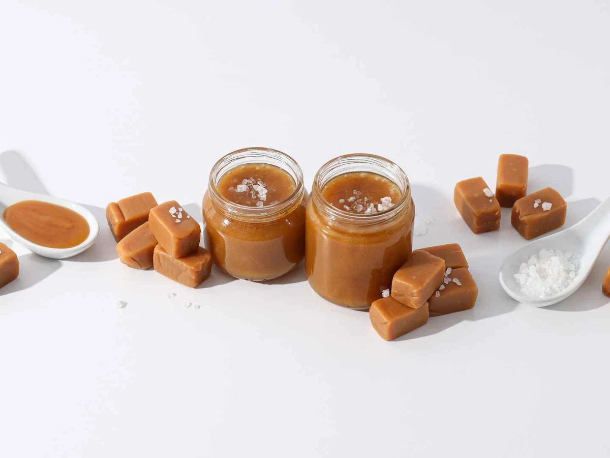 Caramel cubes, two small caramel jars, spoons, and scattered salt on white.
