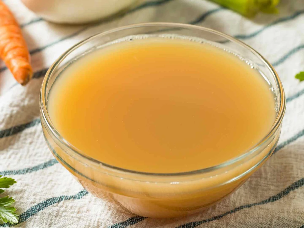 A clear bowl of golden chicken broth beside carrots and parsley
