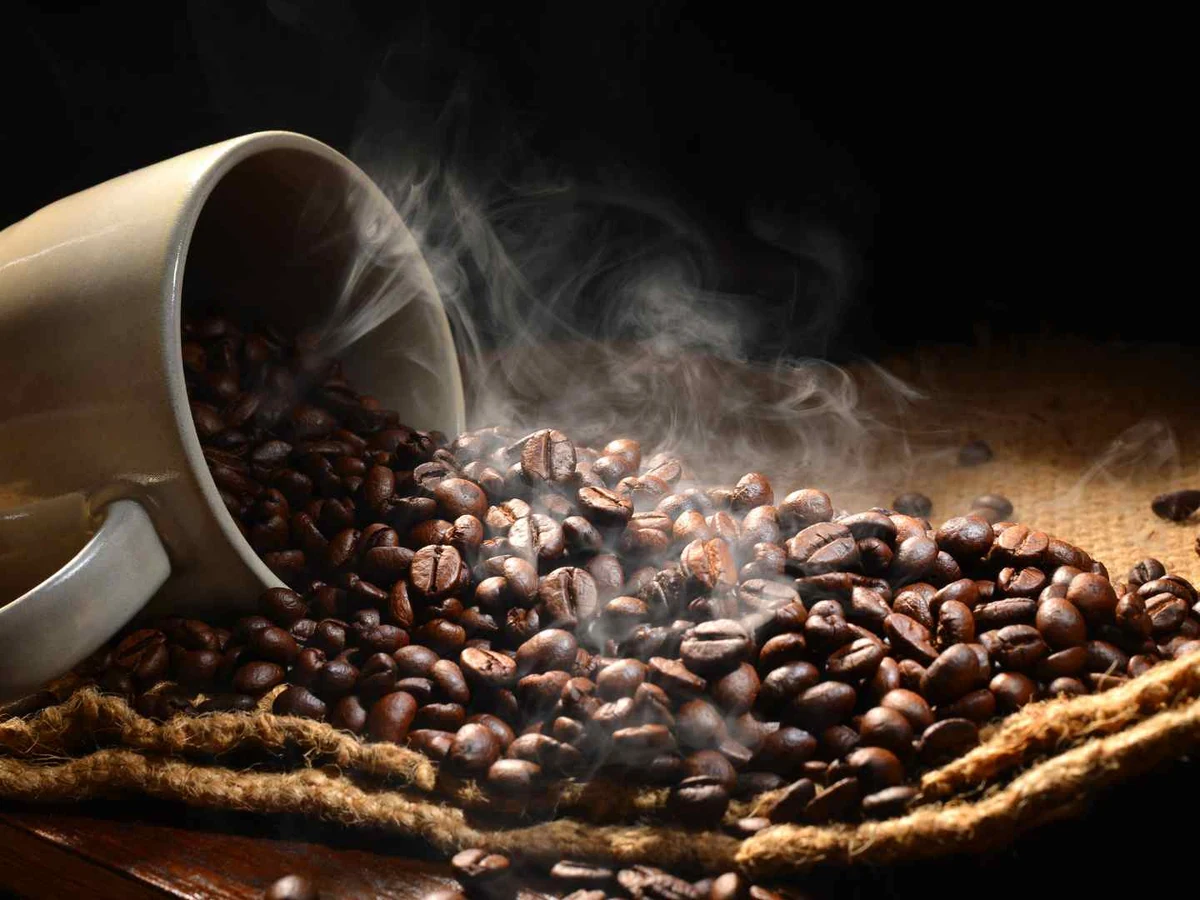 Roasted coffee beans spilling from a tipped cup with visible steam.