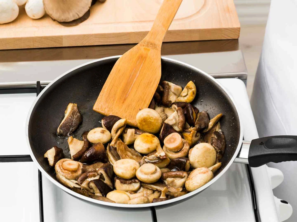 Mixed mushrooms cooking in a frying pan while a person stirs them.