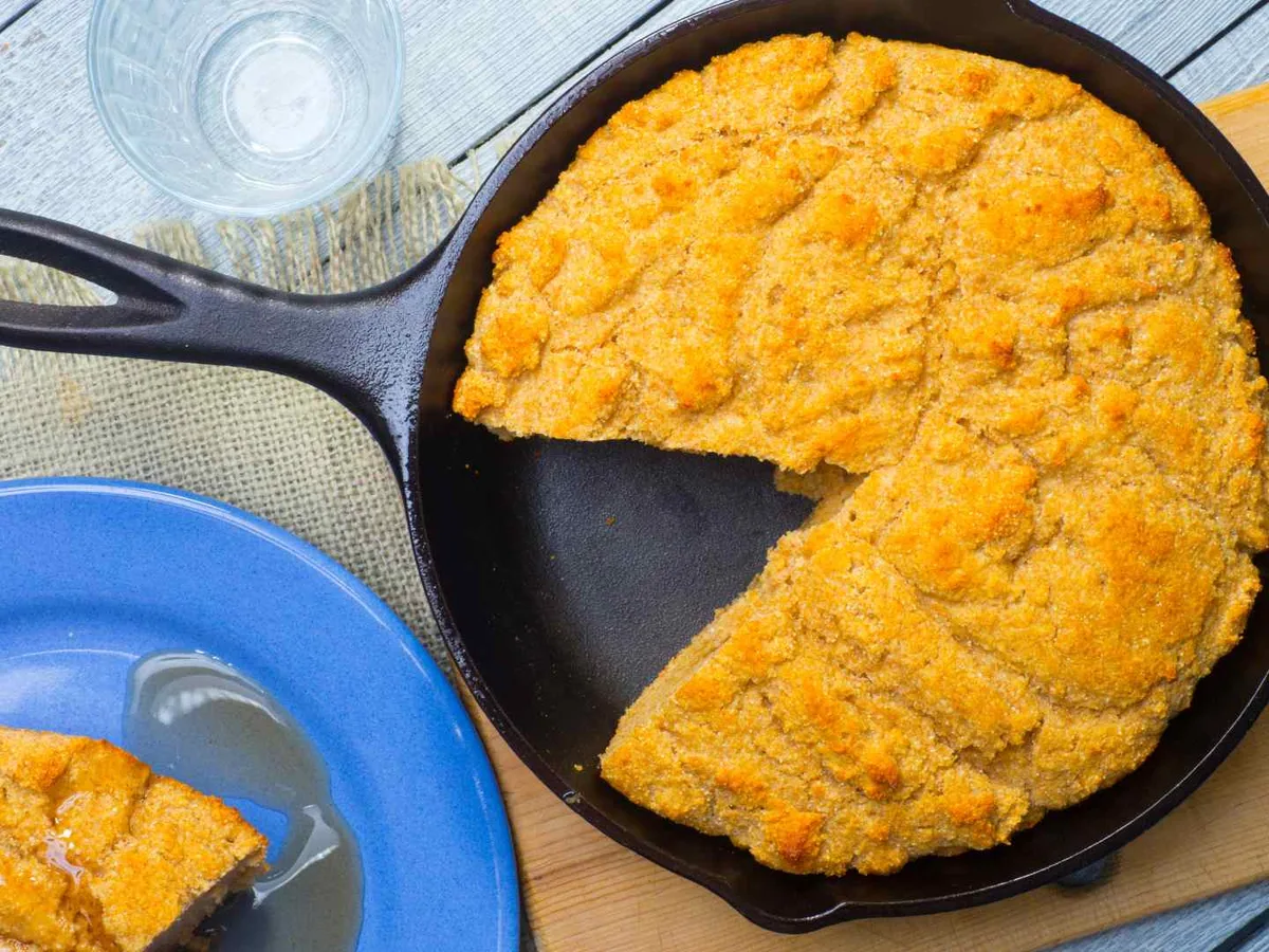A round cornbread in a cast-iron skillet with one wedge removed.