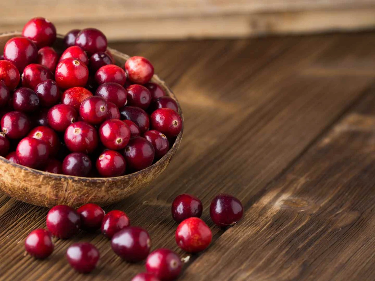 Fresh cranberries in a wooden bowl with loose berries on a table.