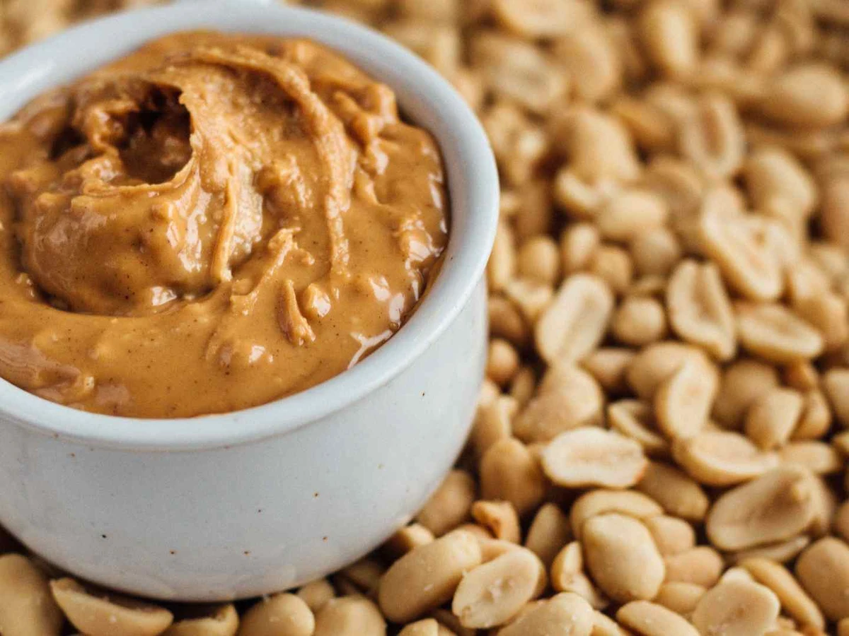 A white ramekin of chunky peanut butter surrounded by shelled peanuts.