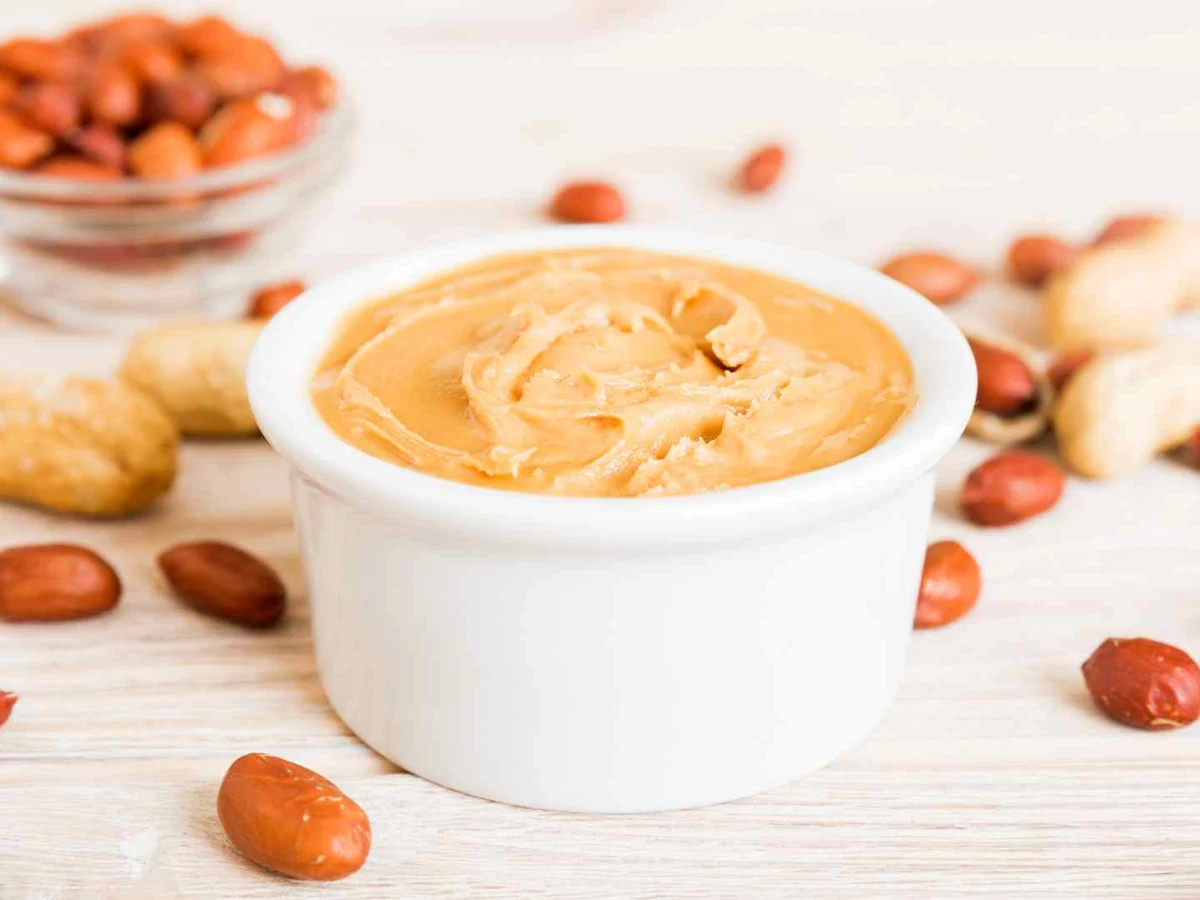 A white ramekin of creamy peanut butter with peanuts scattered nearby.