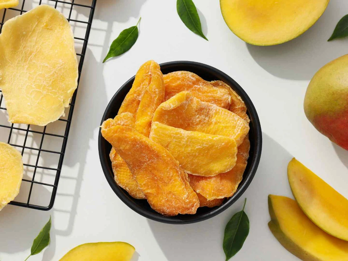 Dried mango slices arranged around fresh mangoes and green leaves.