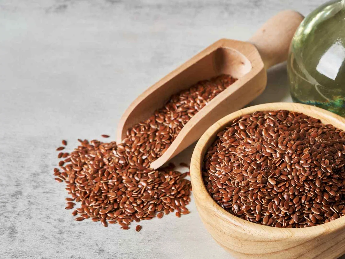 A bowl and scoop of flaxseed beside a bottle of oil