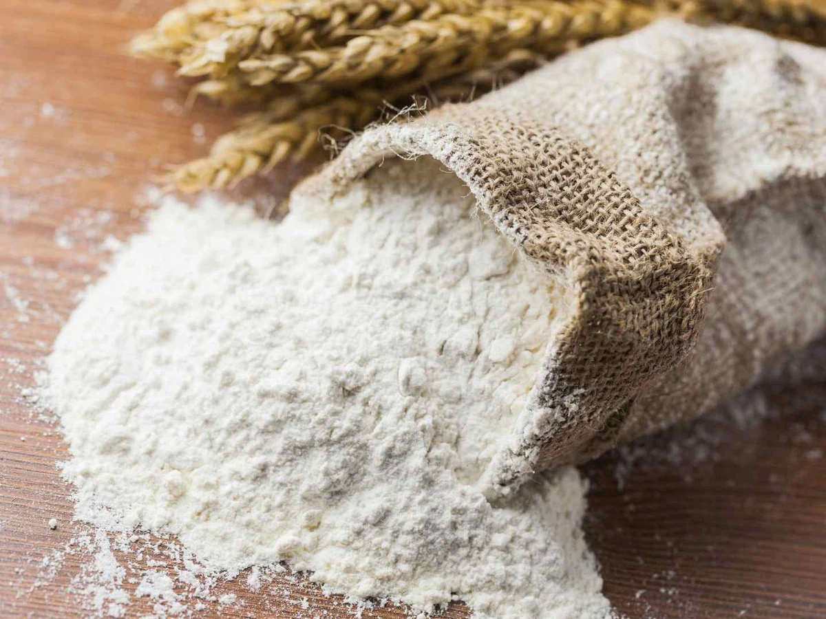 A tipped burlap sack spilling white flour across a wooden table.