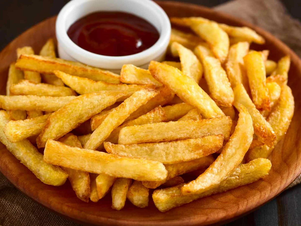 Plain French fries on a wooden plate beside a small cup of ketchup.