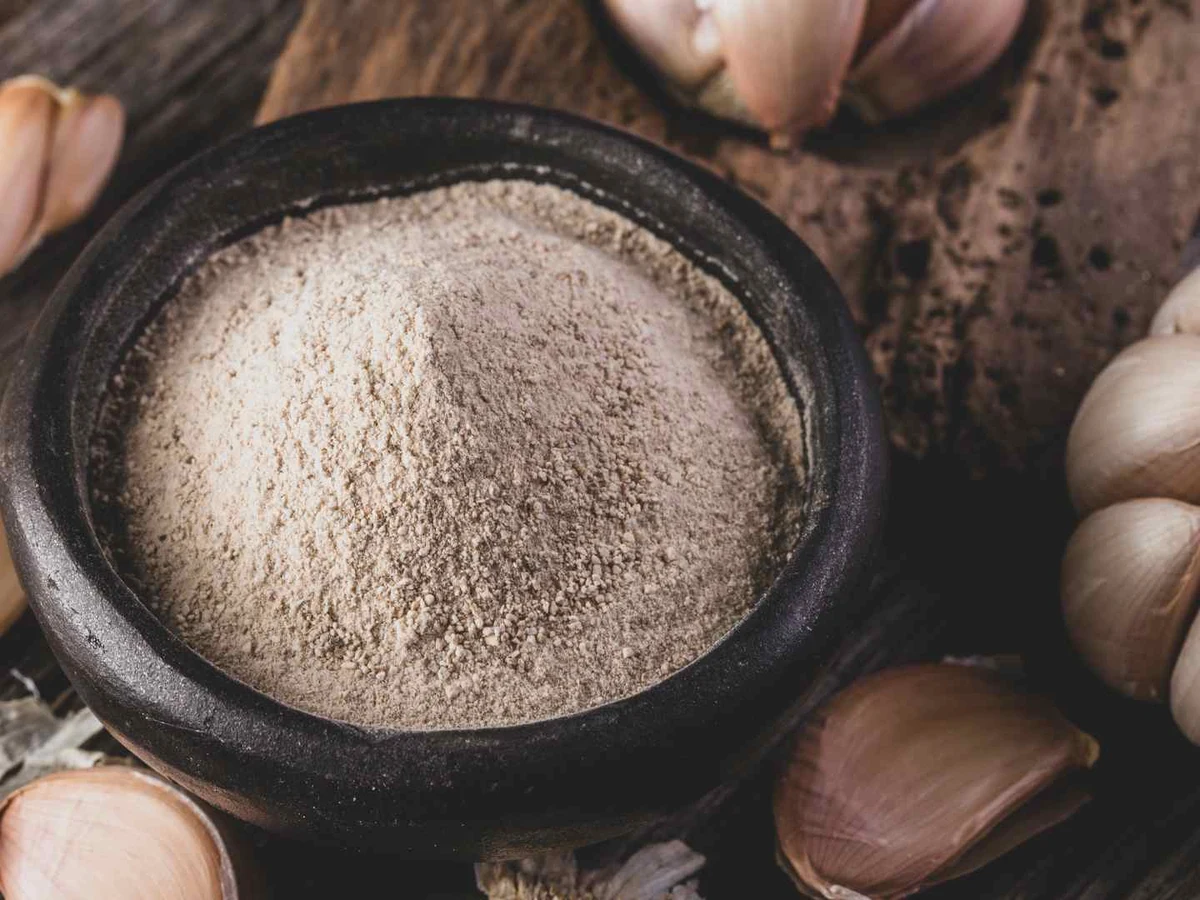 Garlic powder mounded on a spoon above a dark bowl with garlic cloves.