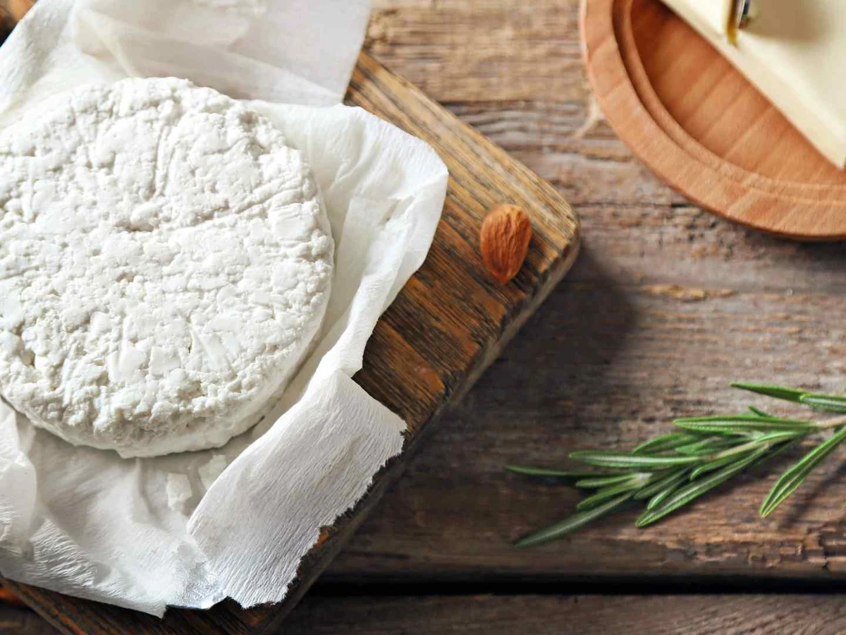 A white goat cheese round on paper with almonds and rosemary nearby.