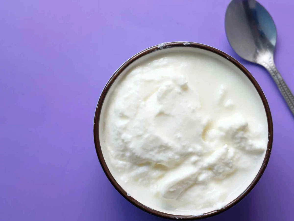 White Greek yogurt in a dark-rimmed bowl beside a metal spoon on a purple surface.