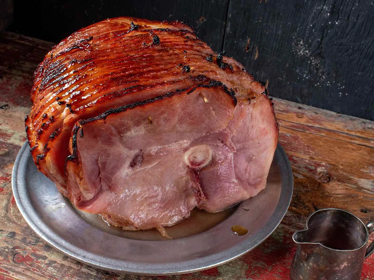 A glazed whole ham on a metal platter beside a small pitcher