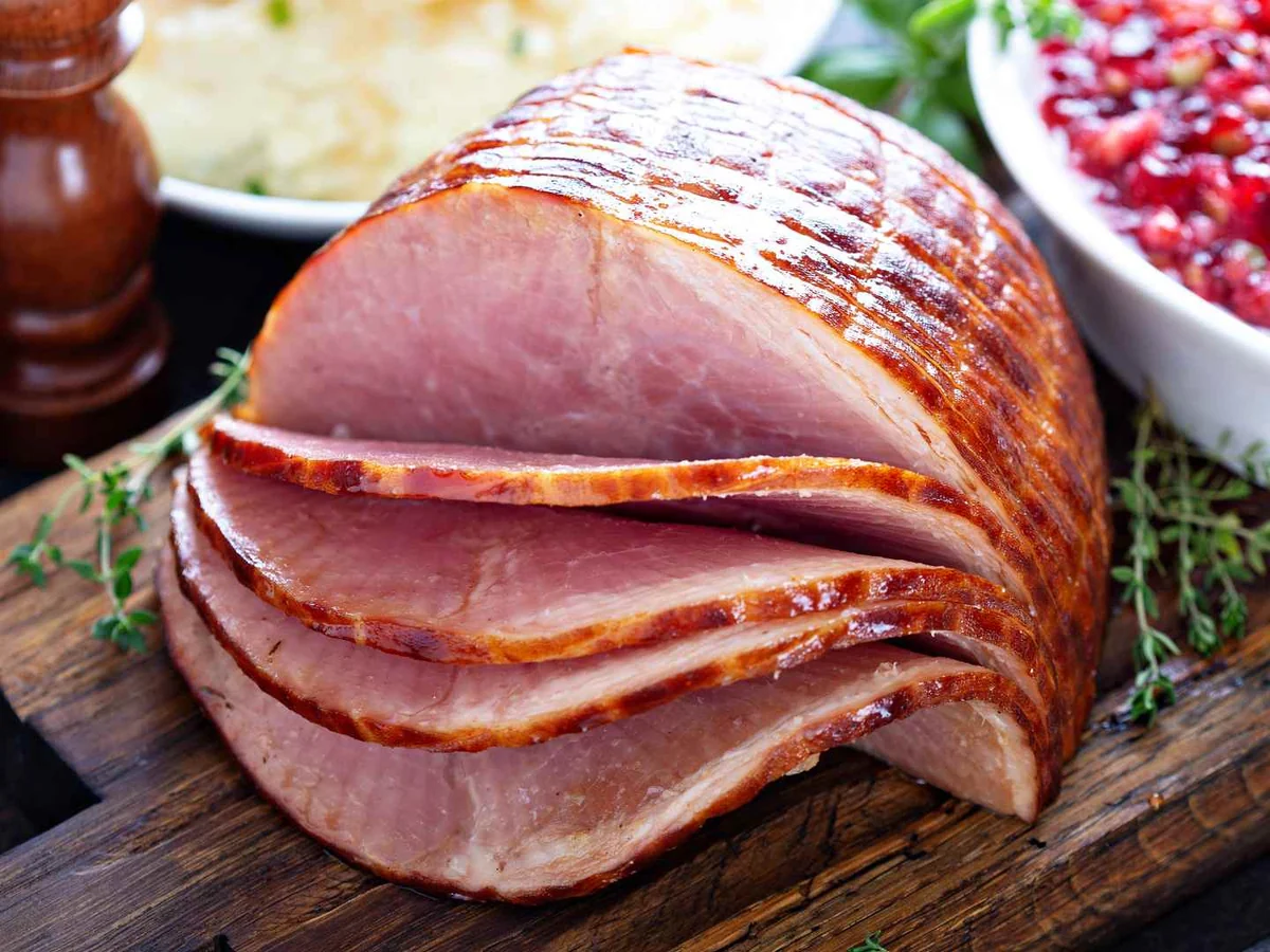 A sliced glazed ham on a wooden board with side dishes behind it