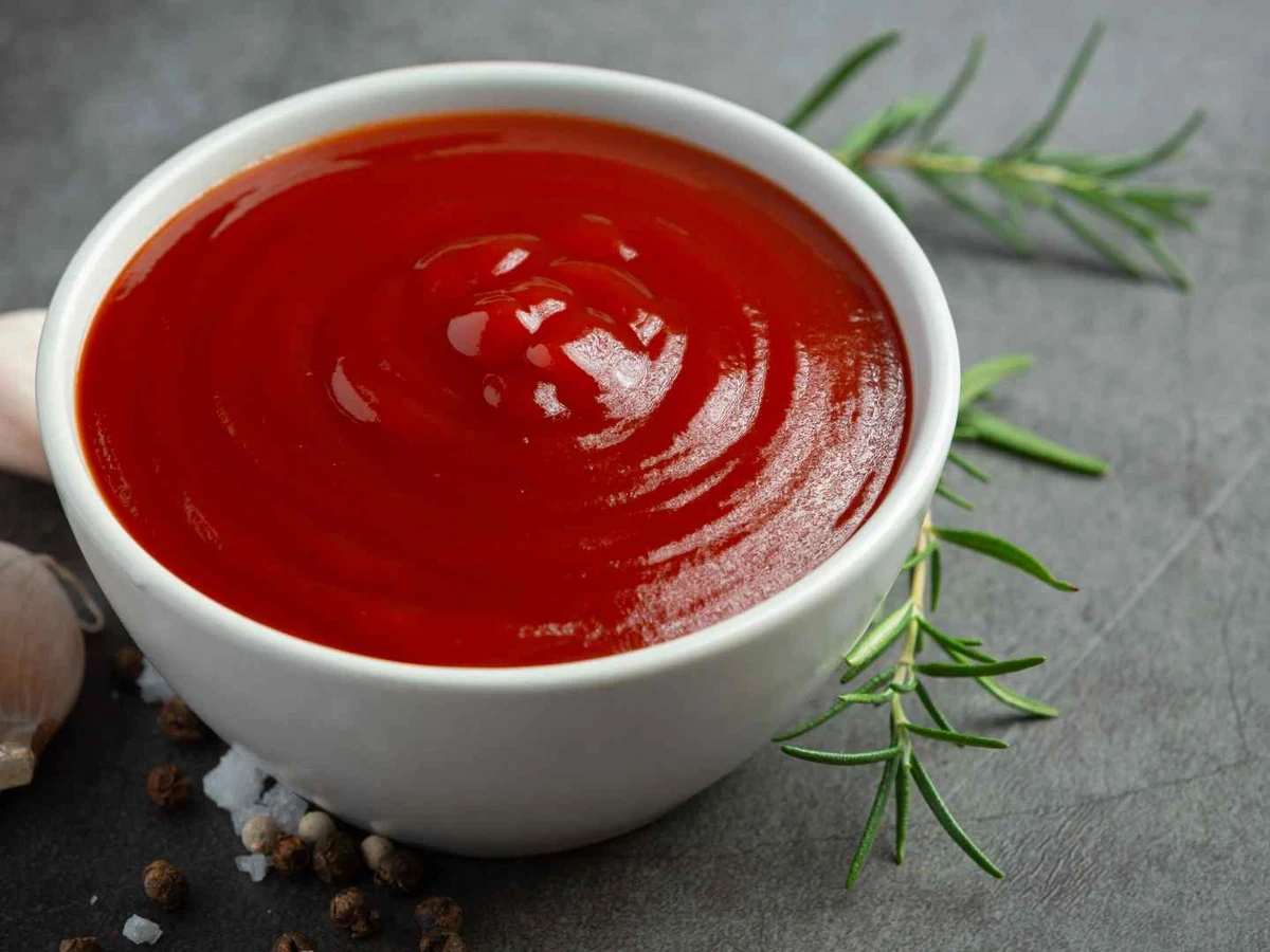 Red ketchup in a white bowl beside garlic cloves and rosemary.