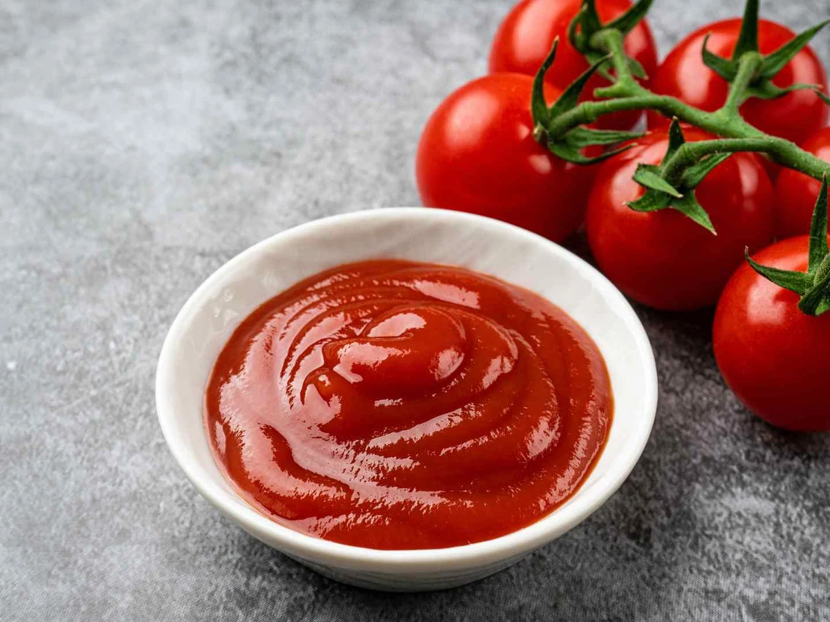 Red ketchup in a white bowl beside tomatoes on the vine.