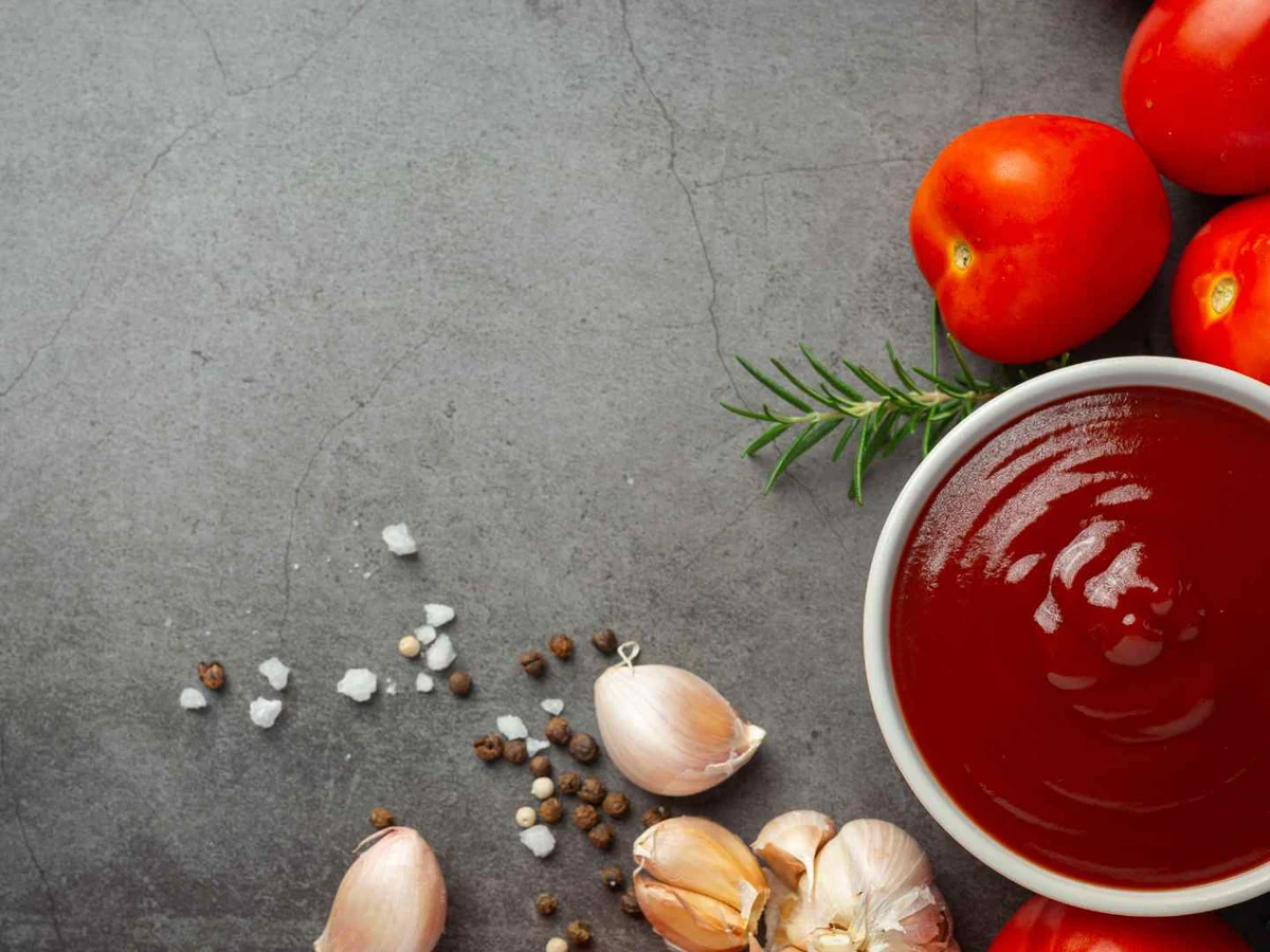 Red ketchup in a bowl beside tomatoes, garlic, herbs, salt, and peppercorns.
