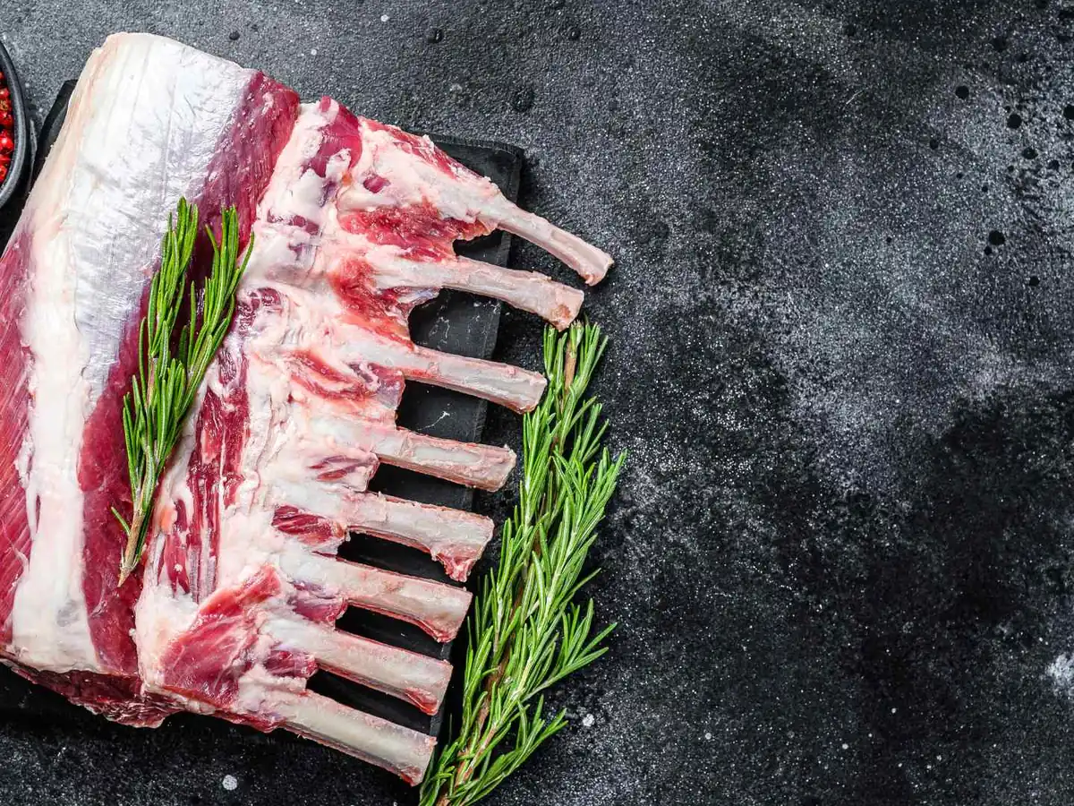 A raw rack of lamb on a dark board with rosemary and peppercorns