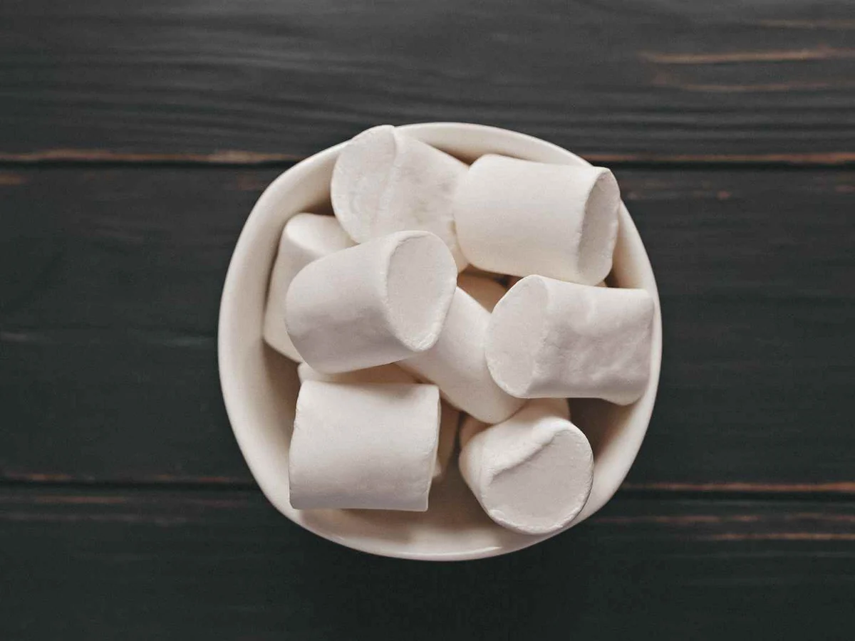 A white bowl of large white marshmallows on a dark wooden table