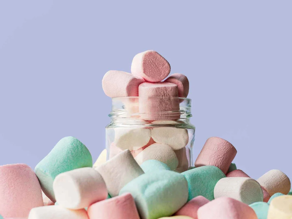 Pastel marshmallows spilling around a glass jar against a pale purple background
