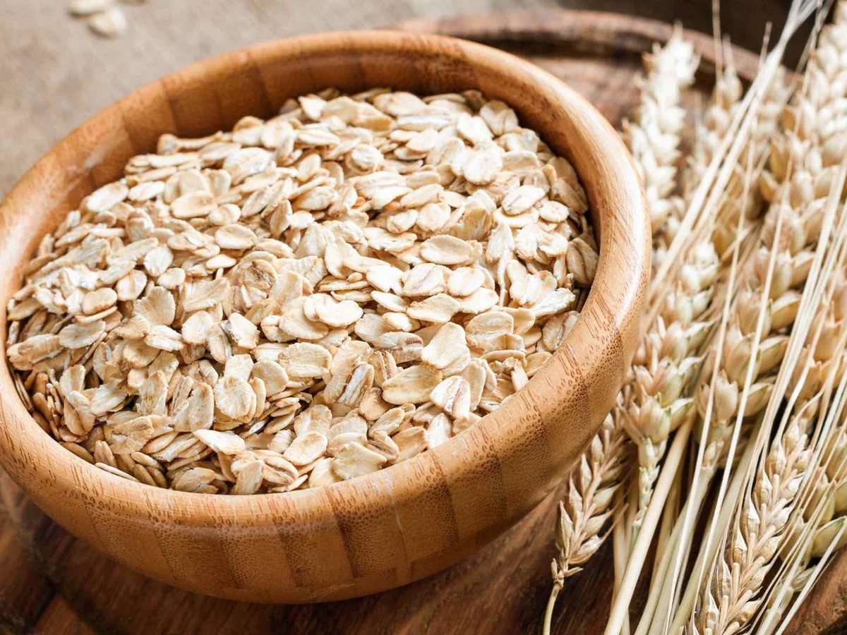 Rolled oats spill from a wooden bowl beside standing wheat stalks.