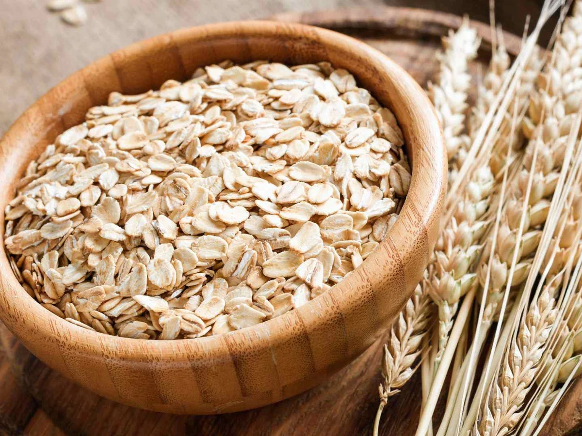 A bowl of rolled oats beside wheat stalks on a blue-gray surface.