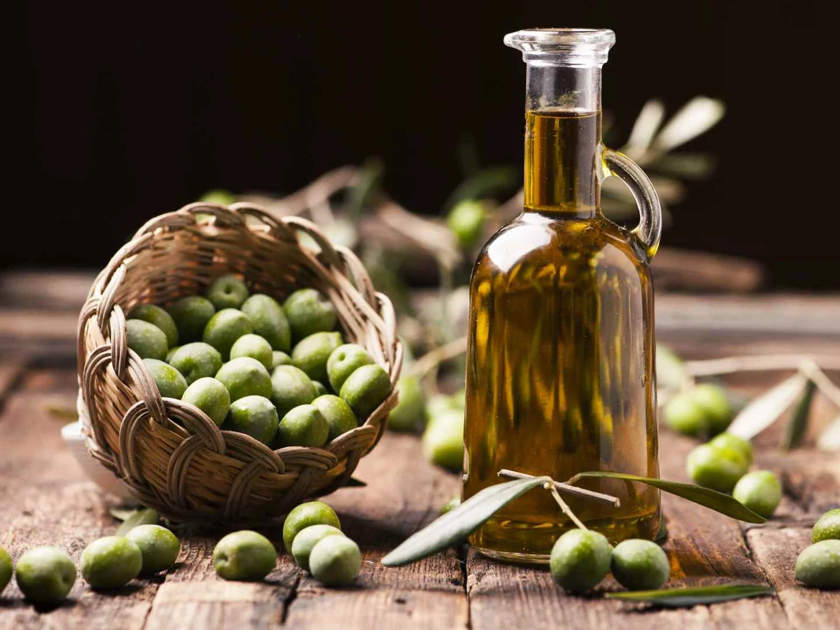 A bottle of olive oil beside a basket of green olives and loose olives.