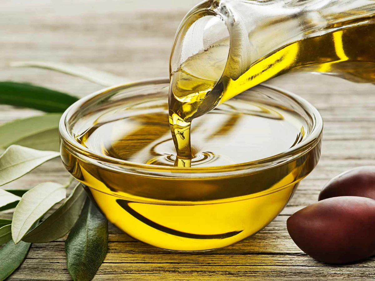 Olive oil pouring into a clear glass bowl beside leaves and dark olives.