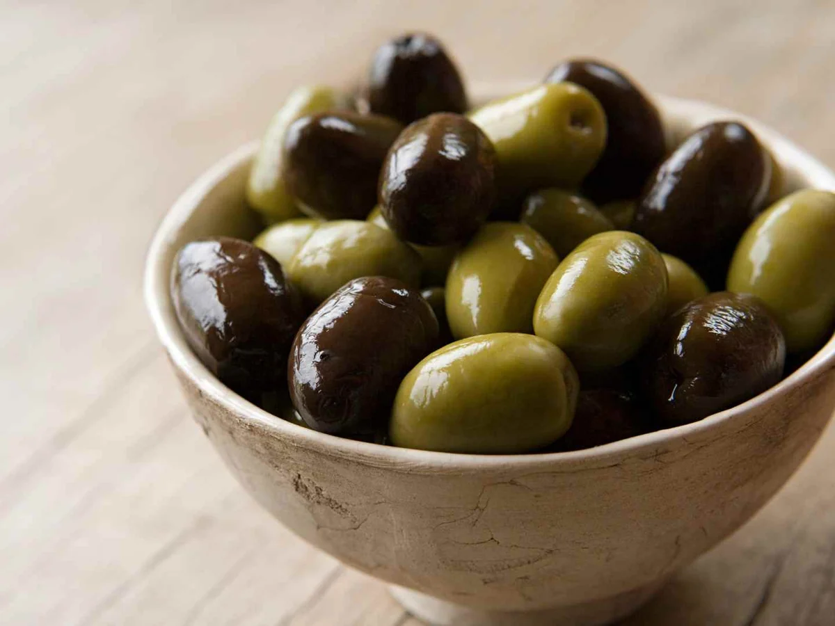 A ceramic bowl filled with green and dark purple olives on a light table