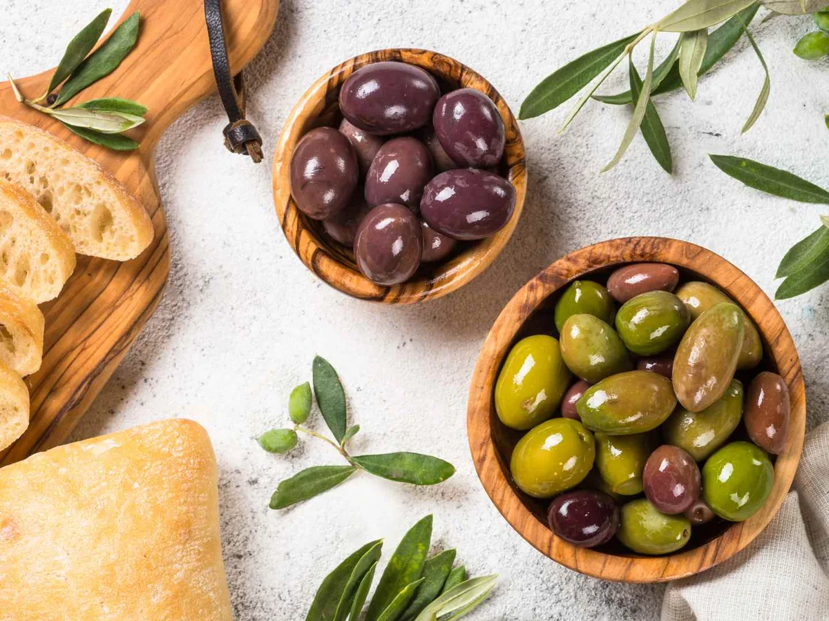 Two wooden bowls of mixed olives beside bread, cheese, and olive branches